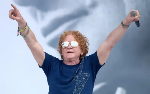 Wokalista Simply Red z uniesionymi rękami, trzymający mikrofon. Ma charakterystyczne rude włosy i okulary przeciwsłoneczne.