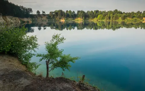 Spokojne jezioro wapienne z odbijającymi się w wodzie drzewami i niebem. Brzeg porośnięty zielenią, z widocznymi skalnymi zboczami.