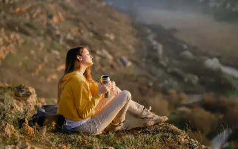 A young woman enjoys the art of doing nothing, holding a thermos and basking in the sun on a rocky hillside.