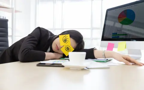 En kvinna sover vid sitt skrivbord med post-it-lappar som föreställer ögon på ögonlocken. Det är uppenbart att hon tycker det är tråkigt på jobbet.