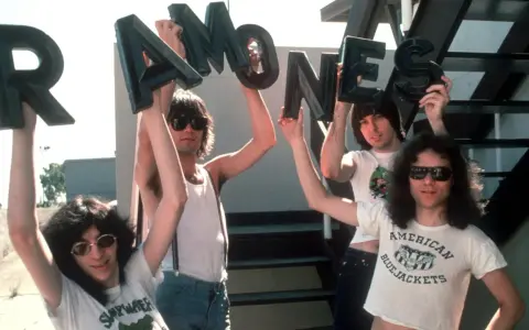 Vier Männer halten die Buchstaben des **titels von Ramones** hoch. Sie tragen Sonnenbrillen und Band-T-Shirts.