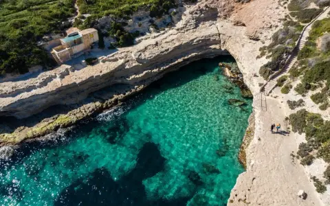 Malta plaże: turkusowa woda w zatoce, skaliste wybrzeże i mały domek. Dwie osoby spacerują ścieżką.