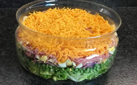 Une salade 7 étages colorée dans un grand bol en verre, avec des couches de laitue, petits pois, oignons rouges, jambon, œufs durs et fromage râpé.