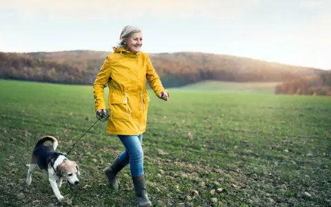 Glad kvinna i gul jacka njuter av en promenad med sin hund i ett fält. En härlig promenad som bränner kalorier.