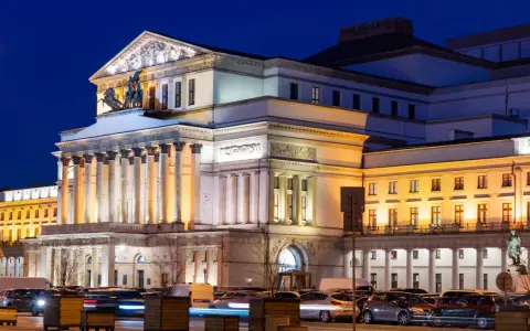 Teatr Wielki w Warszawie, najstarszy teatr w Polsce, nocą. Monumentalna fasada z kolumnami i rzeźbami, oświetlona ciepłym światłem, z samochodami w ruchu ulicznym przed budynkiem.