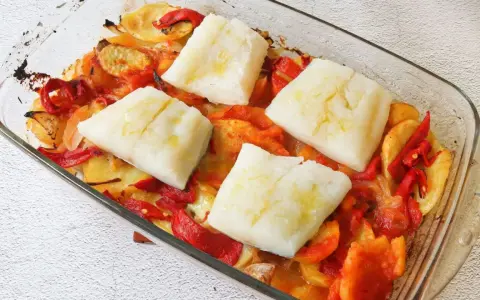 Cuatro porciones de bacalao al horno reposan sobre patatas y pimientos asados en una fuente de vidrio.