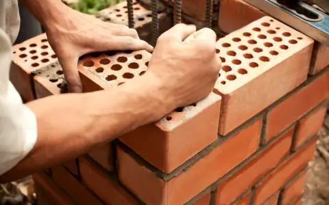 Manos colocan ladrillo con huecos, mostrando sus dimensiones. Construcción de pared con mortero.