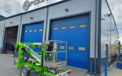 Zielona podnośnik koszowy Nifty 120 stoi przed budynkiem z niebieskimi bramami.