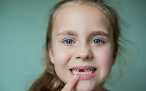 Uśmiechnięta dziewczynka z niebieskimi oczami pokazuje palcem lukę po wypadającym zębie.