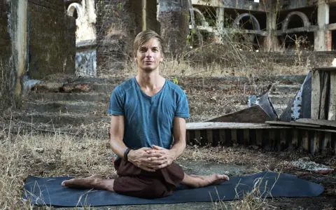 Man i blå tröja sitter i lotusställning på en yogamatta i ruiner. En stunds natthiko meditation i en övergiven miljö.
