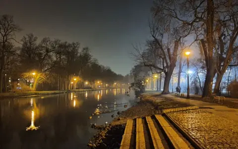 Mglisty wieczór nad **Bydgoszczą**, latarnie oświetlają spokojną **rzekę** i aleję drzew.