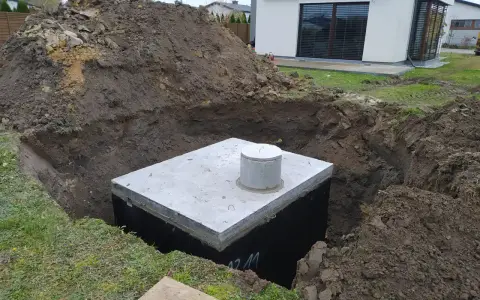 Gotowy odpływ z rynny do ziemi, betonowy zbiornik w wykopie, obok hałda ziemi i nowoczesny dom.