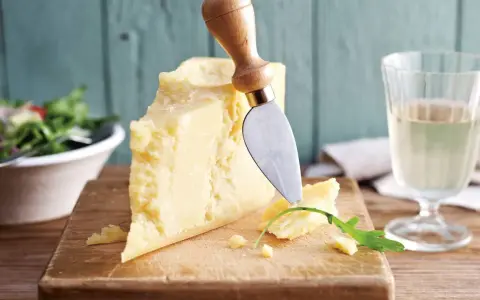 Ein Stück Parmesan, perfekt für vegetarische Gerichte, wird mit einem Käsemesser angeschnitten. Daneben ein Glas Weißwein und ein Salat.