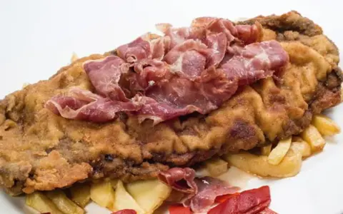 Cachopo asturiano rebozado con jamón serrano encima, acompañado de patatas fritas y pimientos rojos.