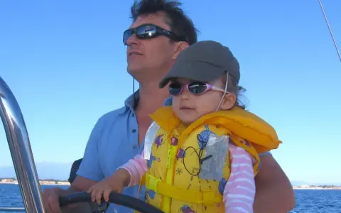 Un homme, le chef de bord bateau, tient son enfant sur ses genoux, naviguant sous un ciel bleu.