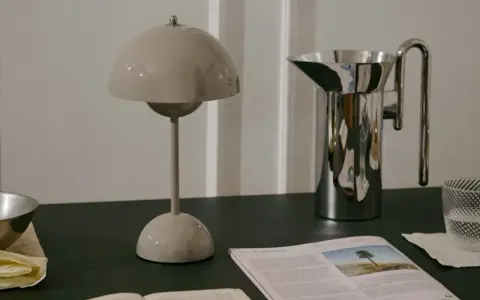 En beige lampa i studio lawahl-stil står bredvid en blank vattenkanna. En tidning med ett träd på omslaget ligger framför.