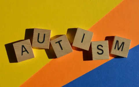 Lettere di legno formano la parola AUTISM su uno sfondo colorato. La domanda "autistici si nasce o si diventa" risuona.