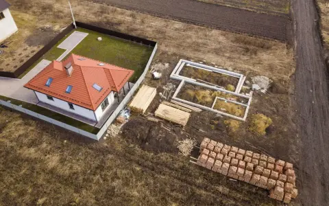 Zabudowa zagrodowa w budowie: gotowy dom z czerwonym dachem, fundamenty, drewno i palety cegieł na placu budowy.