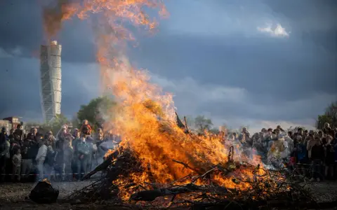 Stort bål med flammor som slår upp mot en dramatisk himmel. En folkmassa samlas för att bevittna ett traditionellt valborg firande.