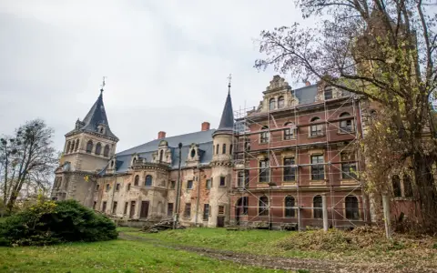 Pałac w Krowiarkach, majestatyczna budowla z wieżami i ceglaną fasadą, otoczony jesienną zielenią, częściowo w rusztowaniach.