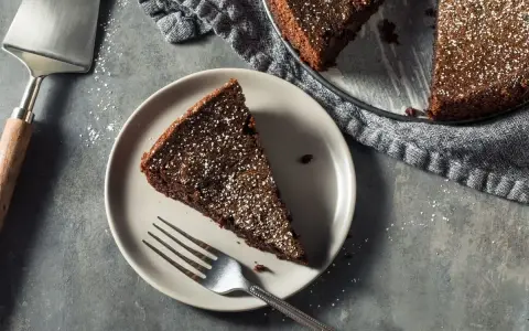 Una fetta di torta all'acqua al cacao, spolverata di zucchero a velo, su un piatto con una forchetta. Accanto, la torta intera e una paletta.