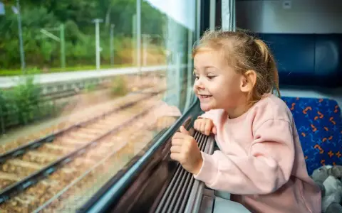 Uśmiechnięta dziewczynka patrzy przez okno pociągu. Zastanawia się, od ilu lat można jeździć samemu pociągiem, marząc o podróżach.