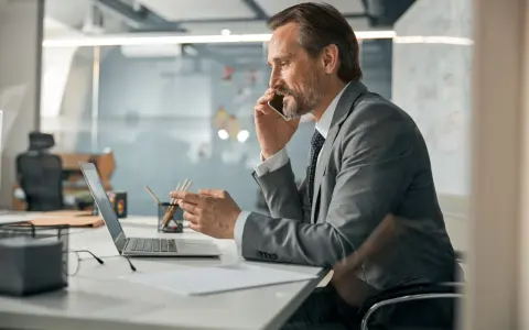 Biznesmen w garniturze rozmawia przez telefon, pracując przy laptopie. To typowy dzień w życiu CEO, co to znaczy być liderem.