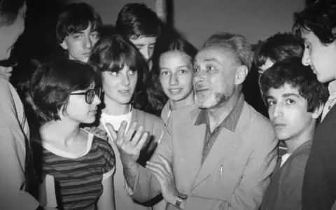 Un uomo anziano con barba e capelli grigi parla animatamente a un gruppo di giovani. Se questo è un uomo riassunto, è un uomo che ispira e coinvolge.