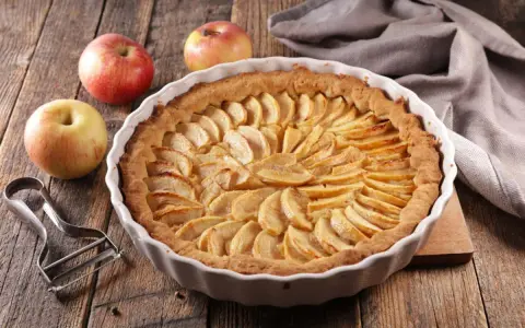 Torta di mele umida e soffice, con mele fresche e un pelapatate su un tavolo di legno rustico.