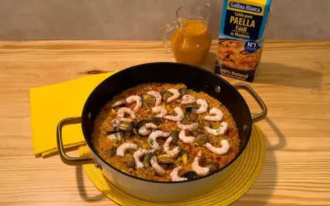 Delicioso arroz a la marinera en paellera, con gambas y mejillones. Al lado, un cartón de caldo para paella de marisco y una jarra con líquido naranja.