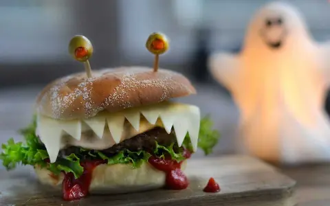 En läskig halloween hamburgare med osttänder och olivögon. En spöklampa lyser upp i bakgrunden.