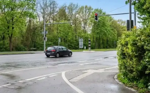 Ein dunkles Auto fährt auf nasser Straße an einer roten Ampel vorbei, neben der ein ampel blitzer steht.