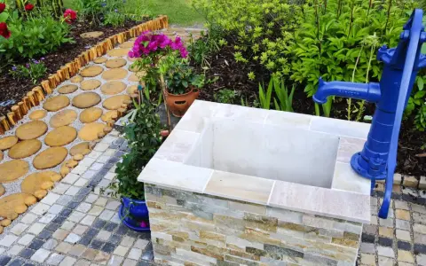 Ein Gartenweg führt zu einem frisch gebauten Brunnen mit blauer Handpumpe.