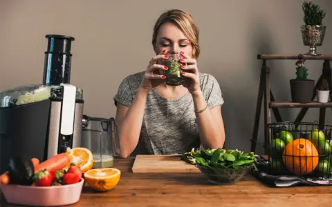 Kvinna dricker grön juice, omgiven av färsk frukt och grönsaker. Hon verkar njuta av sin hälsosamma dryck, kanske som en del av hur deffar man.