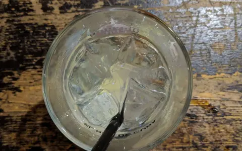 Ein Glas mit Eiswürfeln und einem schwarzen Strohhalm, bereit für einen erfrischenden Gin Tonic auf rustikalem Holztisch.