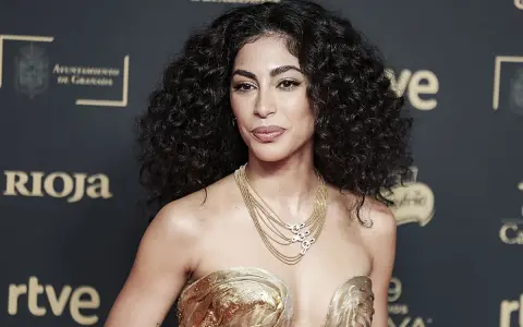Mujer con espectacular pelo rizado, peinado con volumen y brillo. Luce un vestido dorado y un collar de cadenas.