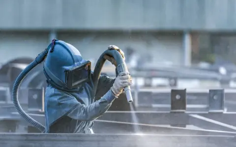 Pracownik w masce piaskuje metalowe elementy. To część pracy z maszynami do metalu, gdzie precyzja jest kluczowa.