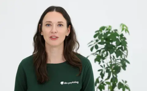 En kvinna med mörkt hår och blå ögon, klädd i en grön tröja med texten "din psykolog", talar. Bredvid henne står en grön växt. Hon ger dig verktyg att hantera sorg.