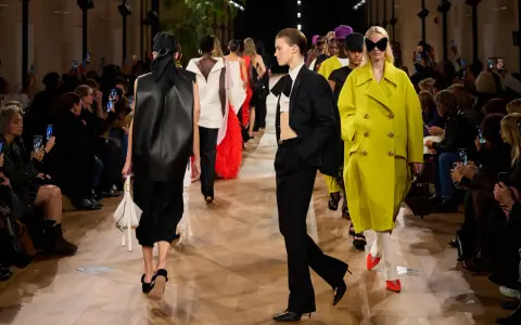 Sfilata Balenciaga: modelli con abiti audaci, tra cui un cappotto giallo acceso e un completo nero, sotto gli occhi del pubblico.
