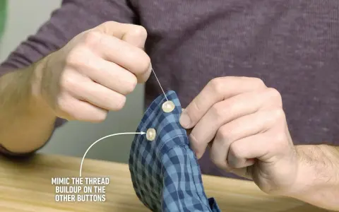 Main tenant une chemise à carreaux bleus, il est en train de coudre un bouton avec une aiguille et du fil blanc.