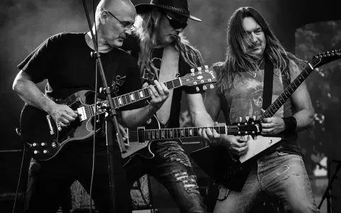 Trzech gitarzystów grających na scenie, przypominających klimatem ballady rockowe lat 80.