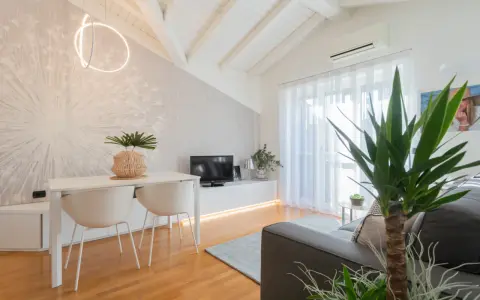 Luminoso appartamento mansardato con travi a vista, zona giorno con tavolo, sedie, TV e divano. Parete con carta da parati decorativa.