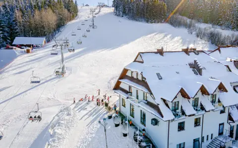 Hotel ze stokiem narciarskim dla dzieci. Kolejka krzesełkowa wwozi narciarzy na ośnieżony stok, a obok hotelu bawią się dzieci.