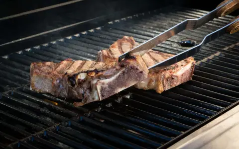Zwei saftige Cowboy-Steaks werden mit einer Zange auf einem heißen Grill gewendet. Rauch steigt auf, während die Flammen darunter lodern.