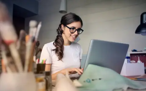 Uśmiechnięta kobieta w okularach pracuje przy laptopie, analizując koszty pracodawcy związane z umową zlecenie.