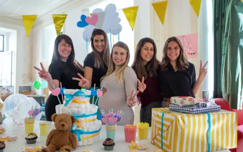 Pięć uśmiechniętych kobiet świętuje baby shower. Na stole tort z pieluch, prezenty i słodkości. To radosne wydarzenie, co to jest baby shower, pełne miłości i oczekiwania.