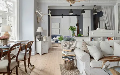 Un salotto con un'atmosfera da arredamento francese chic. Tavolo da pranzo in legno, divano bianco accogliente, lampade e decorazioni rustiche.