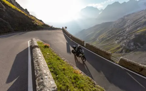 Motocyklista przemierza serpentyny górskiej drogi, odkrywając najpiękniejsze trasy motocyklowe w Europie. Słońce przebija się przez mgłę, oświetlając majestatyczne szczyty.