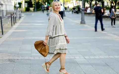 Kobieta w sukience w geometryczne wzory i espadrylach na koturnie idzie ulicą z brązową torebką.