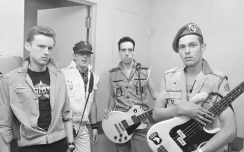 Les membres de The Clash posent avec leurs guitares. L'un porte un t-shirt "The Clash".